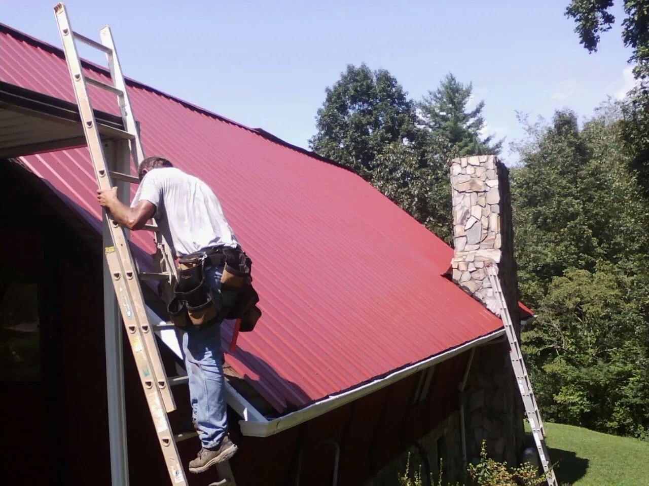 Roofer on a ladder beside a red metal roof and stone chimney — Soffit & Fascia Repair by Roofing Brothers in Collingswood